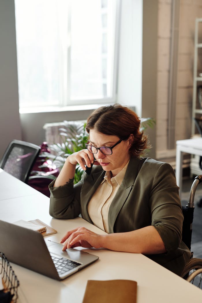 Businesswoman in a wheelchair working from home office on a laptop, focused on a phone call.